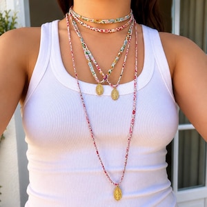 Peut inclure: Un ensemble de colliers superposés composé de trois cordons en tissu à motifs floraux colorés. Deux cordons sont plus courts et présentent de petits pendentifs ovales dorés, tandis que le plus long porte un pendentif ovale doré unique.