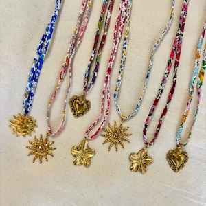 Peut inclure: Plusieurs colliers avec des cordons en tissu à motifs floraux. Les pendentifs sont dorés, en forme de soleils, de cœurs et de fleurs. Les cordons sont dans des tons de bleu, rose et rouge, avec divers motifs floraux.
