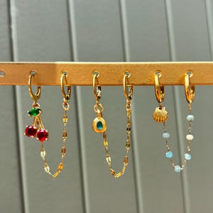Op de afbeelding: Vijf gouden creolen met verschillende bedels. De bedels zijn een kers, een groene edelsteen, een schelp en twee blauwe edelstenen.