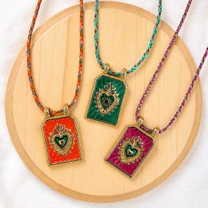 Peut inclure: Trois pendentifs rectangulaires sur des cordons tressés. Chaque pendentif présente un cadre doré, un emblème de cœur sacré central avec une petite pierre sombre et un fond en émail vif orange, vert ou magenta. Les cordons sont multicolores.