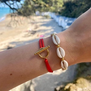 May include: Two bracelets on a wrist. One bracelet is red cord with a gold toggle clasp. The other bracelet is white seashells with gold beads.