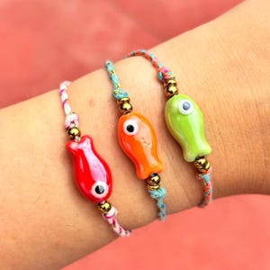 Puede incluir: Tres pulseras de abalorios con forma de pez de colores con cuentas doradas y cuerda blanca, azul y rosa. Los abalorios de pez tienen un solo ojo negro y son rojo, naranja y verde.