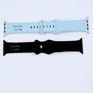 Op de afbeelding: Twee Apple Watch bandjes, een lichtblauw en een zwart, met de tekst "Travel Far, Live Fully" erop gedrukt.
