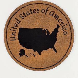 May include: A brown leather coaster featuring a black silhouette of the United States of America with the text "United States of America" around the edge.