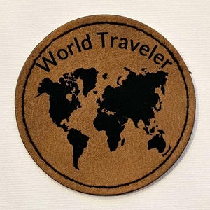 Puede incluir: Un parche de cuero marrón con un contorno negro y un diseño de mapa mundial negro. El texto "World Traveler" está escrito alrededor del mapa.