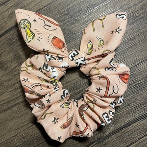 May include: A pink fabric hair scrunchie with a pattern of stars, wands, and other magical objects. The scrunchie is tied in a bow.