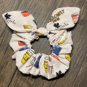 May include: A white scrunchie with a bow, patterned with colorful illustrations of coffee cups, food, and the text "Dragonfly Inn" and "The Girls". The scrunchie is a hair accessory.