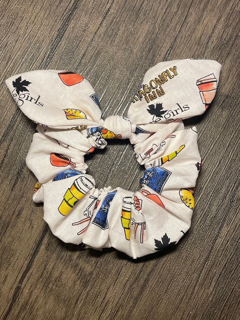 May include: A white scrunchie with a bow, patterned with colorful illustrations of coffee cups, food, and the text "Dragonfly Inn" and "The Girls". The scrunchie is a hair accessory.