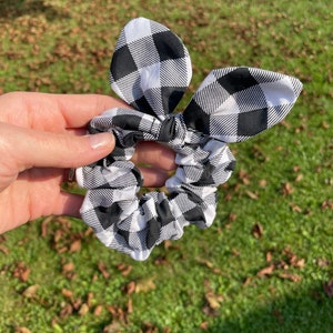 May include: A black and white checkered scrunchie with a bow. The scrunchie is made of fabric and has a gingham pattern. The bow is attached to the scrunchie and is in the shape of bunny ears.
