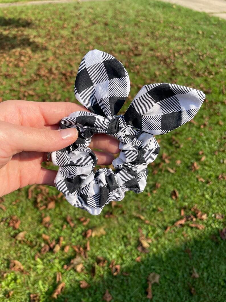 May include: A black and white checkered scrunchie with a bow. The scrunchie is made of fabric and has a gingham pattern. The bow is attached to the scrunchie and is in the shape of bunny ears.