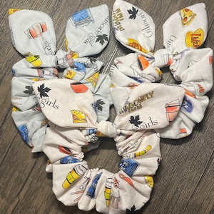 May include: White fabric scrunchies with a repeating pattern of colorful illustrations and text. The illustrations include a coffee cup, a burger, and a television. The text includes the words "Gilmore Girls" and "Dragonfly Inn".