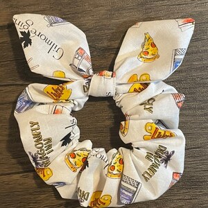 May include: White scrunchie with a bow, featuring a repeating pattern of pizza slices, the Dragonfly Inn, and text from Gilmore Girls. The scrunchie is made of fabric and is designed to hold hair in place.