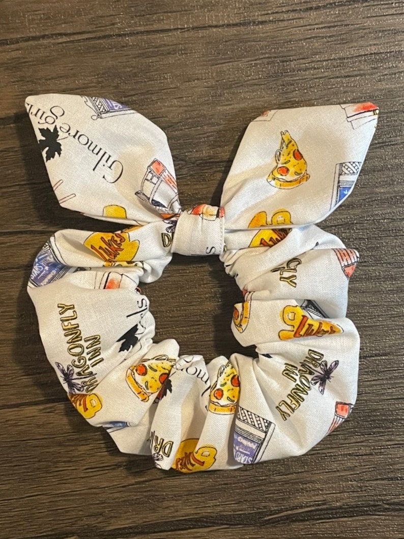 May include: White scrunchie with a bow, featuring a repeating pattern of pizza slices, the Dragonfly Inn, and text from Gilmore Girls. The scrunchie is made of fabric and is designed to hold hair in place.