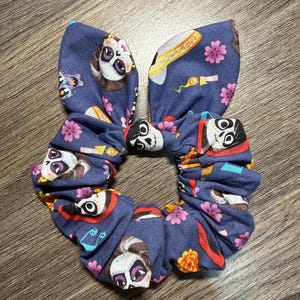 May include: A blue scrunchie with a Day of the Dead design. The fabric is patterned with sugar skull faces, flowers, and festive motifs. The scrunchie is styled with a bow, offering a fun detail to the hair accessory.
