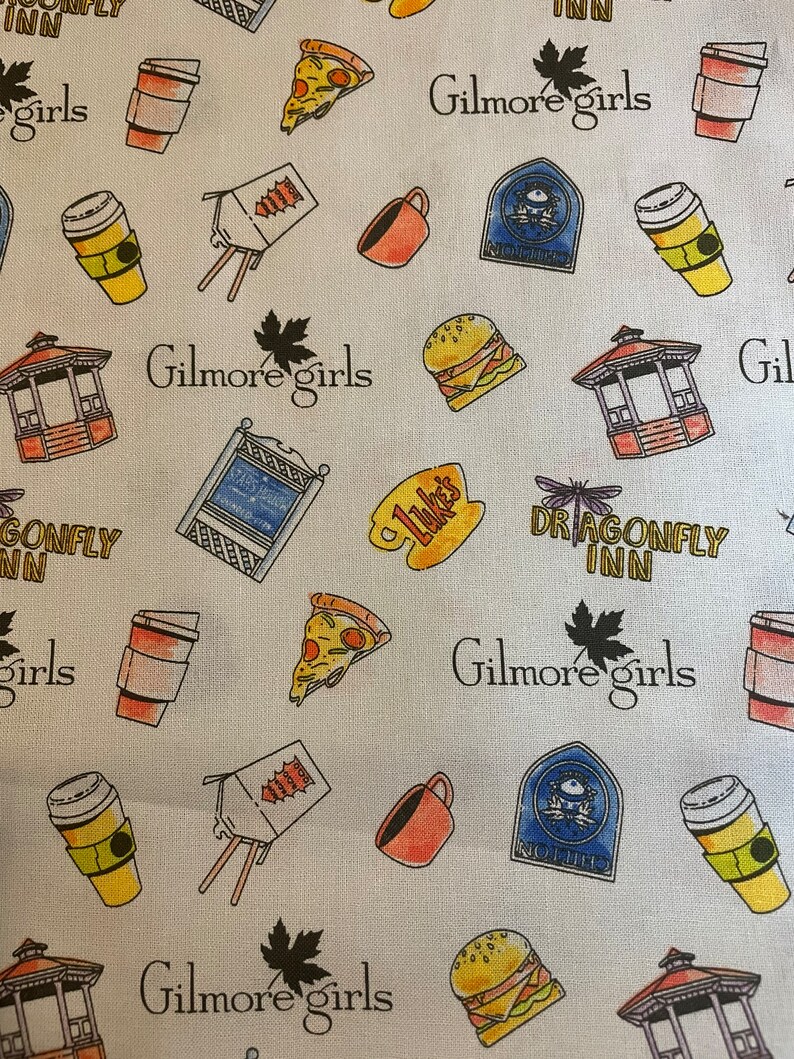 May include: White fabric patterned with various illustrations and text related to the TV show "Gilmore Girls." The design includes coffee cups, pizza slices, burgers, gazebos, and the show's logo. The text includes "Gilmore Girls," "Luke's," and "Dragonfly Inn."