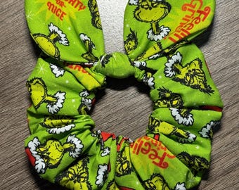Puede incluir: Coletero de tela verde con estampado temático de Grinch. La tela presenta al Grinch con gorro de Papá Noel, copos de nieve y texto que dice "Naughty or Nice" y "Feel Grinchy". El coletero tiene forma de lazo.