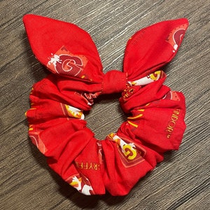 Puede incluir: Scrunchie de pelo de tela roja con un patrón de la casa Gryffindor. El patrón presenta el león de Gryffindor, los colores de la casa y las palabras "Gryffindor" y "Brave".