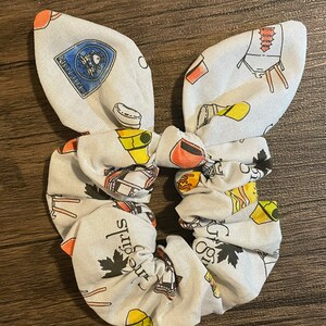 May include: A white scrunchie with a bunny ear bow, patterned with colorful illustrations of cooking tools, maple leaves, and text. The scrunchie is designed for hair accessories.