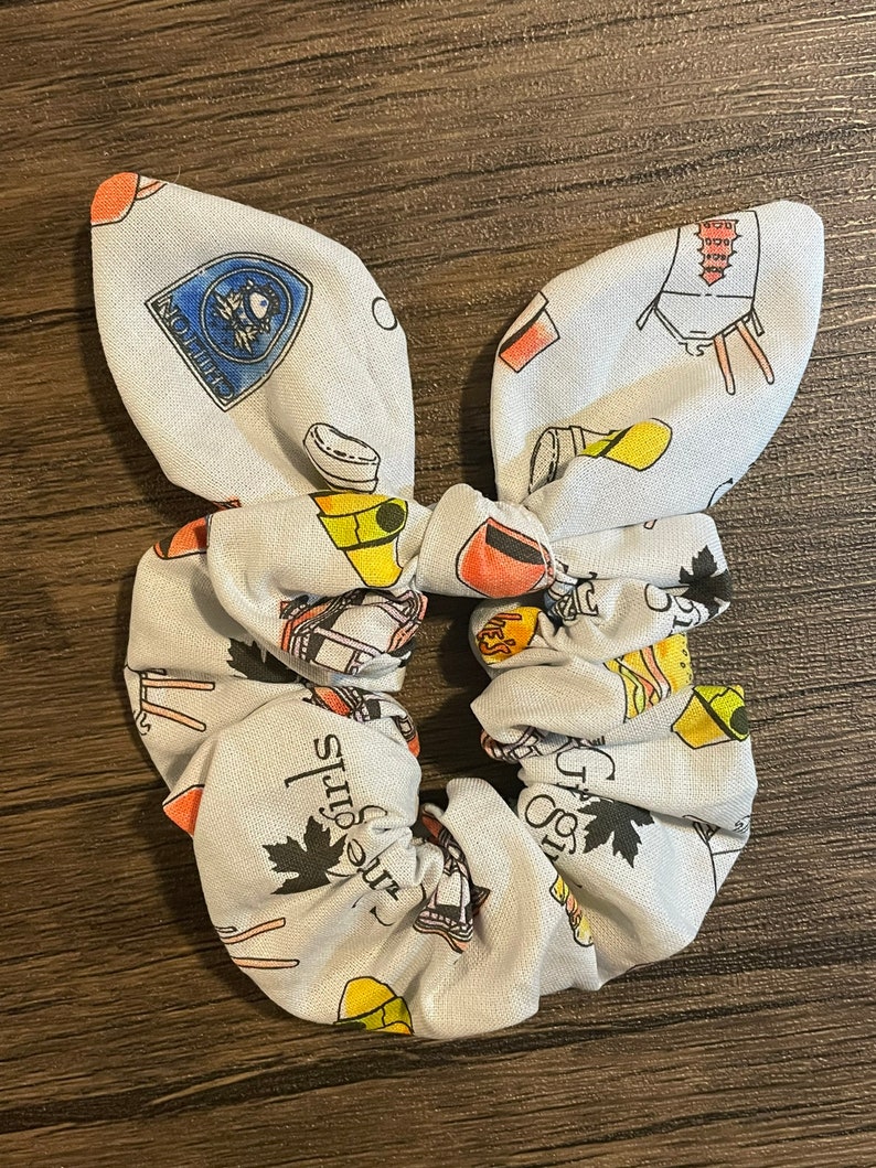 May include: A white scrunchie with a bunny ear bow, patterned with colorful illustrations of cooking tools, maple leaves, and text. The scrunchie is designed for hair accessories.