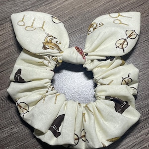 May include: A white fabric scrunchie with a brown and gold print of glasses, wands, and a wizard's hat.