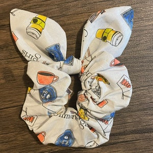 May include: A light blue scrunchie with a bunny ear bow, patterned with coffee cups, books, and the text "Gilmore Girls". The scrunchie is a fun accessory for any hairstyle.