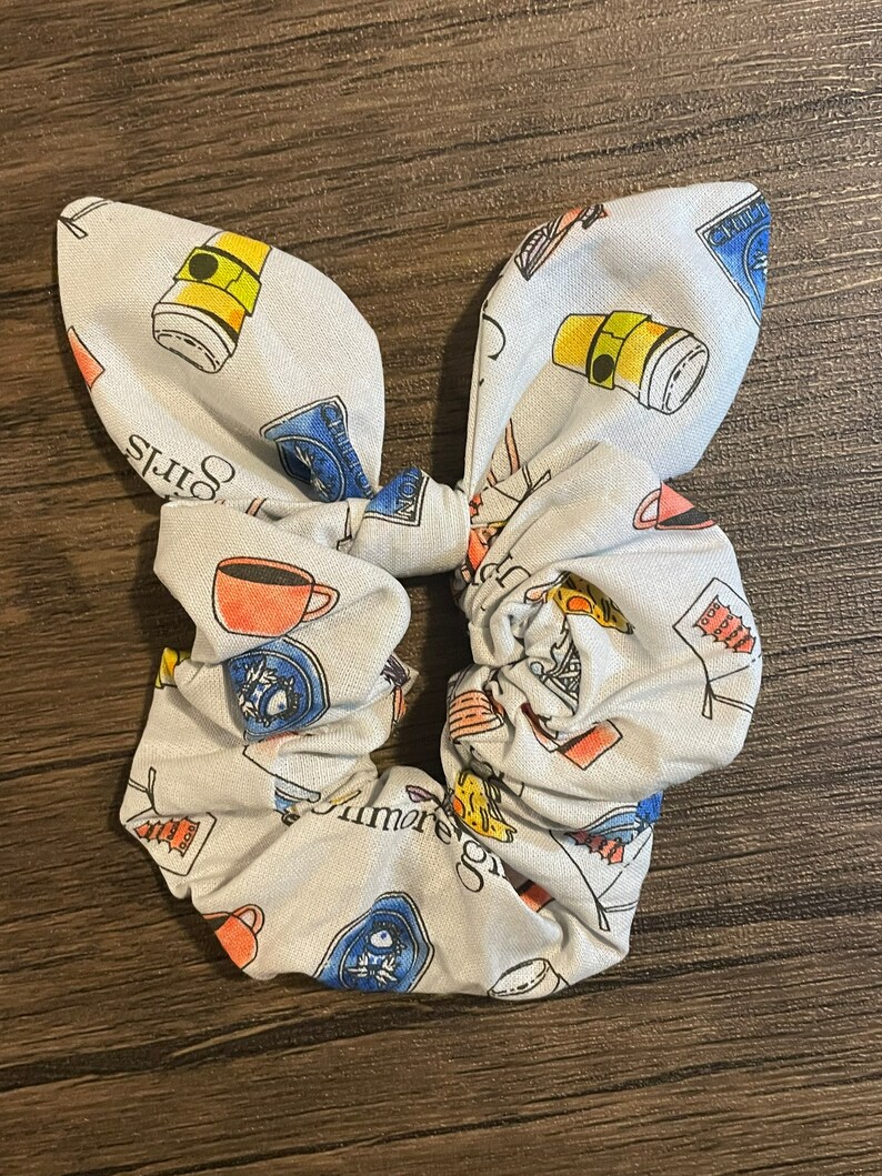 May include: A light blue scrunchie with a bunny ear bow, patterned with coffee cups, books, and the text "Gilmore Girls". The scrunchie is a fun accessory for any hairstyle.
