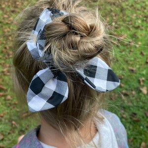 May include: A black and white buffalo plaid bow scrunchie is tied in a bun hairstyle. The scrunchie is made of fabric with a classic checkered pattern. The hair accessory adds a touch of style to the hairstyle.