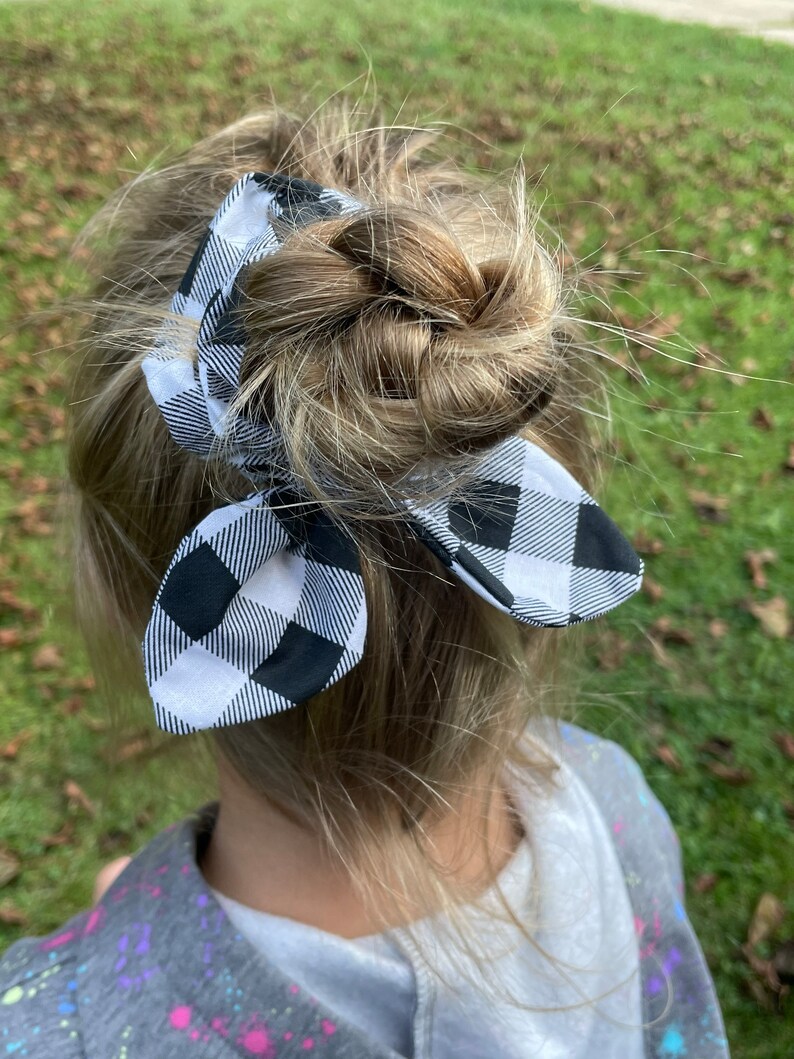 May include: A black and white buffalo plaid bow scrunchie is tied in a bun hairstyle. The scrunchie is made of fabric with a classic checkered pattern. The hair accessory adds a touch of style to the hairstyle.