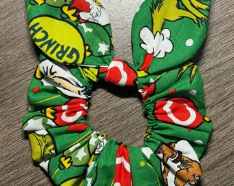 Puede incluir: Coletero de tela verde con estampado temático de El Grinch. El diseño presenta a El Grinch, Papá Noel y otros personajes de la historia. El coletero tiene forma de lazo, con la tela reunida alrededor de una banda elástica.