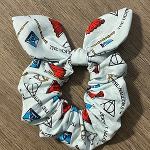 Puede incluir: Un coletero azul claro con fondo blanco y un patrón repetido de símbolos de Harry Potter en rojo y azul, incluyendo el símbolo de las Reliquias de la Muerte, varitas mágicas y las palabras "The Deathly Hallows TM".