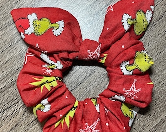 Puede incluir: Scrunchie rojo con fondo blanco con un patrón repetido del Grinch con un gorro de Papá Noel y una estrella blanca.