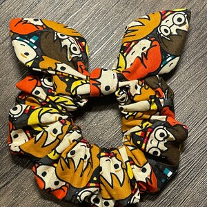 Puede incluir: Un scrunchie de pelo de tela marrón, naranja y amarillo con un estampado de personajes de dibujos animados. Los personajes llevan gafas y tienen el pelo marrón.