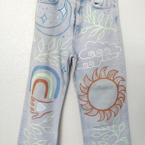 May include: Light blue denim jeans with hand-painted celestial and nature-inspired designs. The artwork includes a crescent moon, stars, sun, rainbow, leaves, and a hand holding a heart. The jeans have a straight leg cut and a frayed hem.
