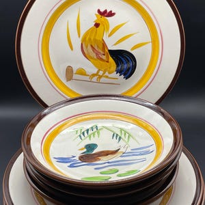 May include: A set of ceramic plates and bowls with hand-painted designs. The large plates feature a rooster, while the bowls display a duck on water. Each piece has a brown rim, yellow and pink accent rings, and a white background.