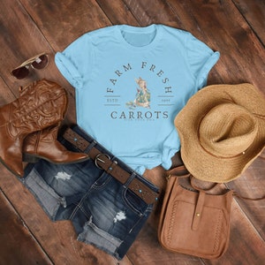 May include: A light blue t-shirt with a graphic of a rabbit holding a carrot. The graphic is surrounded by a circle with the text "Farm Fresh" at the top, "Estd 1901" at the bottom, and "Carrots Pick Your Own" in the center.