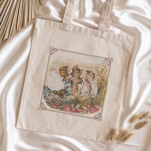 Puede incluir: Una bolsa de tela beige con una ilustración vintage de tres gatitos jugando en un jardín. La ilustración está enmarcada con un borde decorativo.