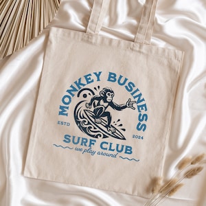 Peut inclure: Sac fourre-tout en toile beige avec texte bleu et un motif de singe surfeur. Le texte indique "MONKEY BUSINESS SURF CLUB" avec "we play around" en dessous. Le motif comprend un singe sur une planche de surf et l'année 2024.
