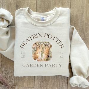 Peut inclure: Un sweat-shirt beige avec un imprimé graphique brun de deux lapins dans un jardin. Le graphique comprend le texte "Beatrix Potter", "ESTD 1901", et "Garden Party".