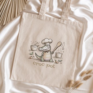May include: A beige canvas tote bag with a cartoon illustration of a crocodile wearing a chef's hat and holding a wooden spoon. The crocodile is surrounded by cooking utensils and ingredients. The text "croc pot" is printed below the illustration.