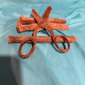 May include: A pair of rusted metal scissors resting on top of three horizontal, weathered, iron railroad spikes. The scissors have two oval finger loops and pointed blades, all showing a textured, oxidized brown surface finish throughout.