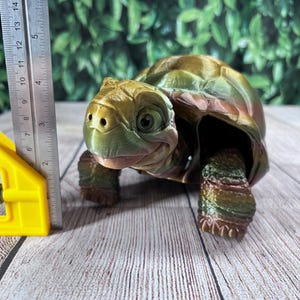 May include: A 3D-printed turtle figurine in green, brown, and gold, with a smiling face. The figurine is on a wooden surface. A ruler shows measurements in centimetres. The turtle is a decorative item.