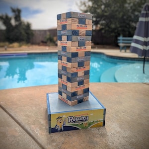 May include: A wooden tower game with blue and brown blocks stacked on top of each other. The blocks have the word "Royalty" printed on them, along with a phone number. The tower is sitting on a yellow box with a cartoon king on it. The box says "Royalty Heating & Air".