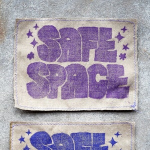 May include: Two beige fabric patches with the words "SAFE SPACE" printed in purple and blue. The patches have a stitched border and are decorated with small star-shaped designs.