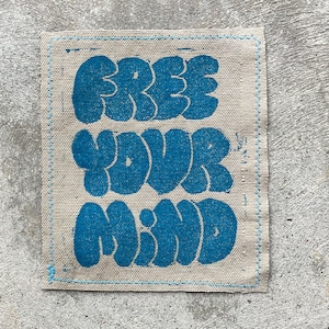 May include: A beige fabric patch with blue lettering that reads "FREE YOUR MIND". The letters are in a bubble font style.