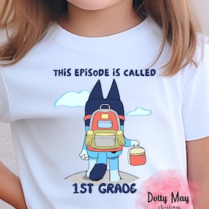 May include: A white t-shirt featuring a cartoon blue dog character wearing a red and gray backpack and carrying a red lunch box. The text above the character reads THIS EPISODE IS CALLED and the text below the character reads 1ST GRADE.