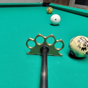 Puede incluir: Un porta bolas de billar de metal dorado con cinco agujeros, una barra de metal negro y tres bolas de billar sobre una mesa de billar de fieltro verde. Una bola es blanca, una es amarilla y una es amarilla con marcas negras.