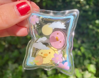 Könnte beinhalten: Ein durchsichtiger Schlüsselanhänger aus Plastik mit einem rosa, gelben und blauen Design. Der Schlüsselanhänger zeigt ein kleines Cartoon-Küken, eine Wolke und eine Sonne. Der Schlüsselanhänger ist an einem rosafarbenen Metallclip befestigt.