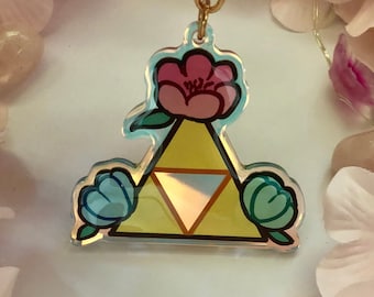 Könnte beinhalten: Ein goldfarbener Schlüsselanhänger mit einem gelben Dreieck und einer rosa Blume oben. Das Dreieck hat eine holografische Oberfläche und ist von zwei grünen Blumen umgeben.