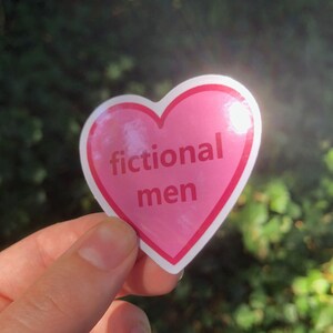 K&ouml;nnte beinhalten: Ein rosa herzf&ouml;rmiger Aufkleber mit wei&szlig;em Rand. Der Aufkleber tr&auml;gt den wei&szlig;en Schriftzug "fictional men". Der Hintergrund ist eine verschwommene Ansicht von gr&uuml;nem Laub.