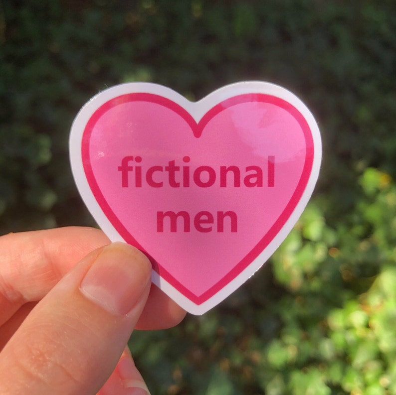 K&ouml;nnte beinhalten: Ein rosa herzf&ouml;rmiger Aufkleber mit den Worten "fictional men" in wei&szlig;em Text. Der Aufkleber hat einen wei&szlig;en Rand und wird vor einem verschwommenen gr&uuml;nen Hintergrund gehalten.